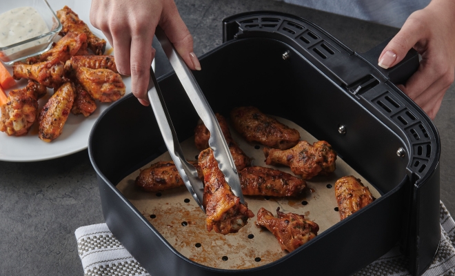 Person removing cooked chicken wings from an air fryer with a liner using tongs