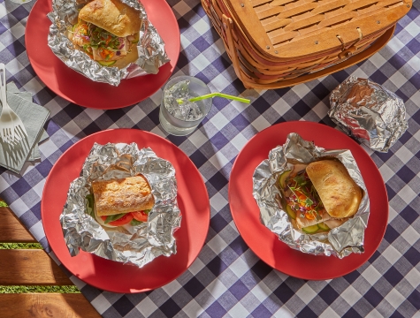 Sandwiches wrapped in aluminum foil sitting on a blanket next to a picnic basket