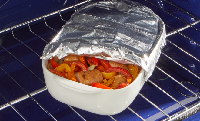 Casserole dish filled with sausage and peppers partially covered with aluminum foil